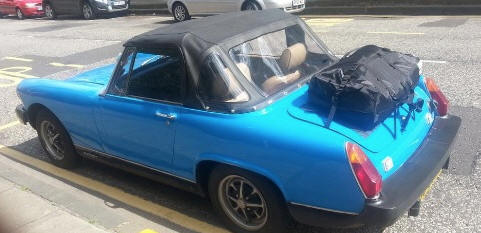 blue rubber bumper late 1970's MG Midget on a residential road in the sunshine hood up with a luggage rack fitted