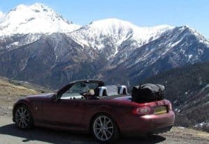 burgandy mazda mx5 with a boot-bag original luggage rack fitted hood down on an alpine pass snowy mountains tops in the background