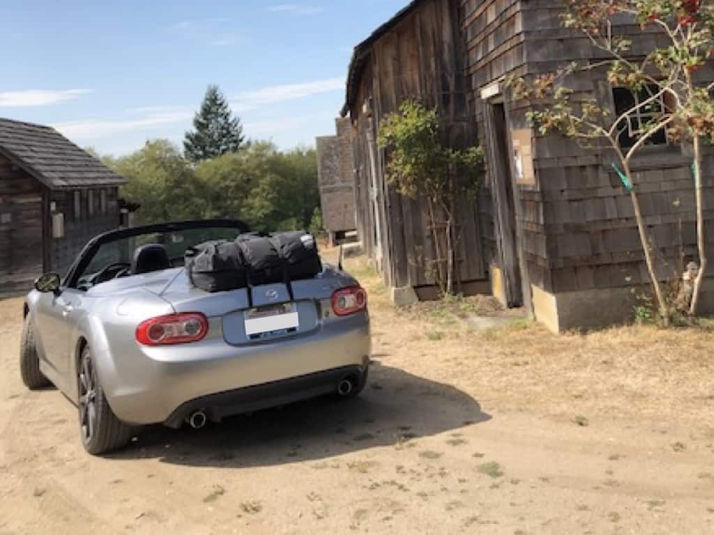 silver mazda miata prht with a boot-bag luggage rack fitted next to a barn